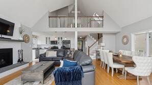 A spacious living room with a fireplace, a dining area with a wooden table and white chairs, and a kitchen in the background. - Coastal Dream Vacation Home at Moody Beach - Sleeps 8 (Wells)