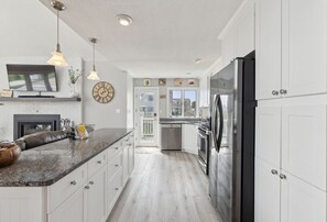 Una cocina moderna con armarios blancos, encimera de granito, electrodomésticos de acero inoxidable, iluminación colgante y un amplio ventanal.