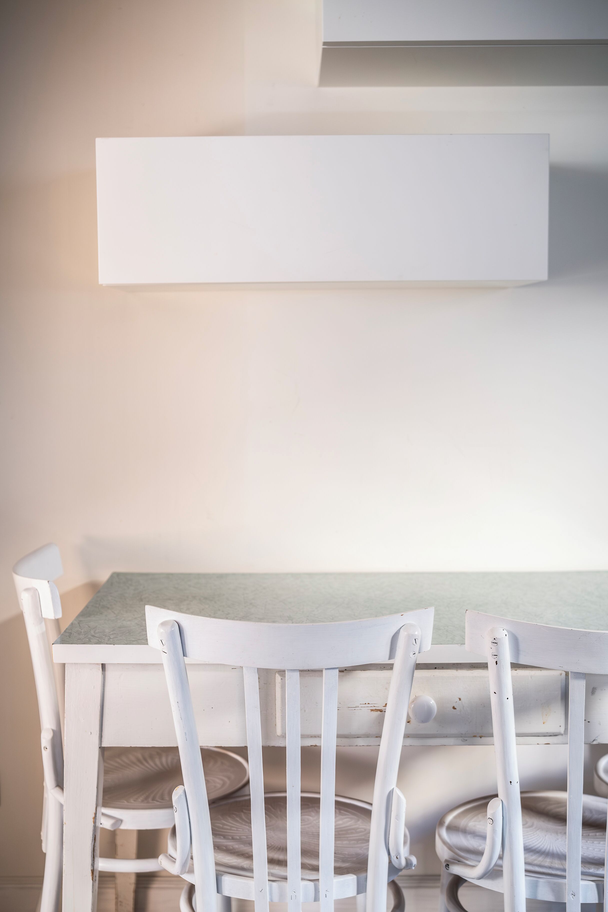 Une table à manger blanche avec des chaises, un mur de couleur claire et une étagère blanche au-dessus.