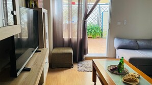 Uma sala de estar moderna com televisão de tela plana, um sofá cinza, uma mesa de centro de madeira e uma porta de vidro deslizante que dá para uma área externa com plantas.