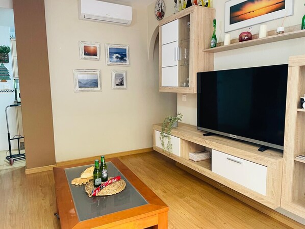 Een moderne woonkamer met een flatscreen televisie, houten kasten, een klein tafeltje met flessen en snacks, en ingelijste foto’s aan de muur.