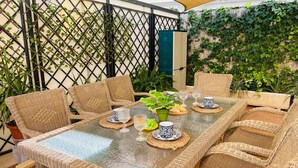 Uma área de refeições ao ar livre coberta, com móveis de vime e uma mesa de vidro, cercada por vegetação e um cercado de bambu.