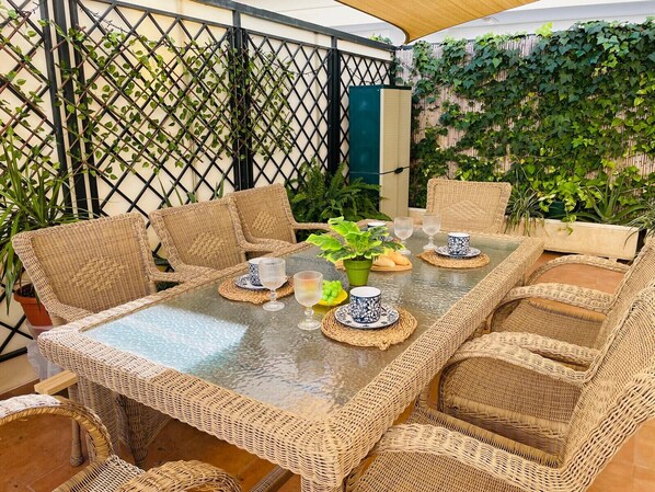 A covered outdoor dining area with wicker furniture and a glass table, surrounded by greenery and a bamboo fence.