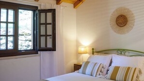 A bedroom with a wooden ceiling, a bed with white and striped bedding, a wall-mounted lamp, and a window with curtains. - Rosemary House (Santiago do Cacém)