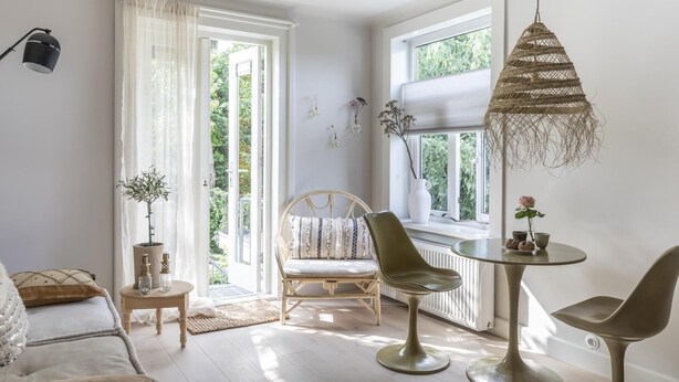 Een lichte kamer met een groot raam, een ronde tafel, twee stoelen, een lamp en een fauteuil met kussen.