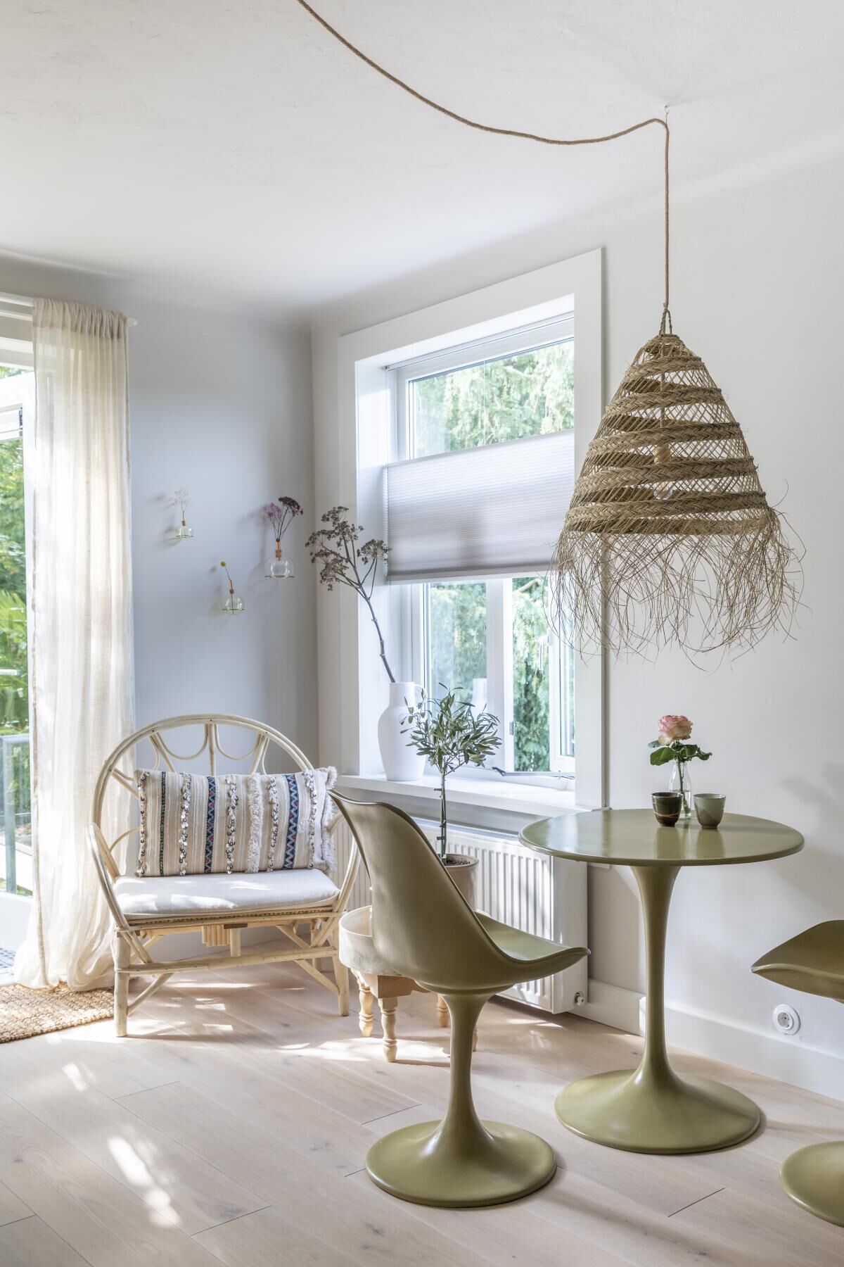 A room with a wicker hanging light fixture, a round table with two chairs, a wicker chair with cushions, and a window with a view of greenery.