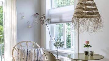 A room with a wicker hanging light fixture, a round table with two chairs, a wicker chair with cushions, and a window with a view of greenery.