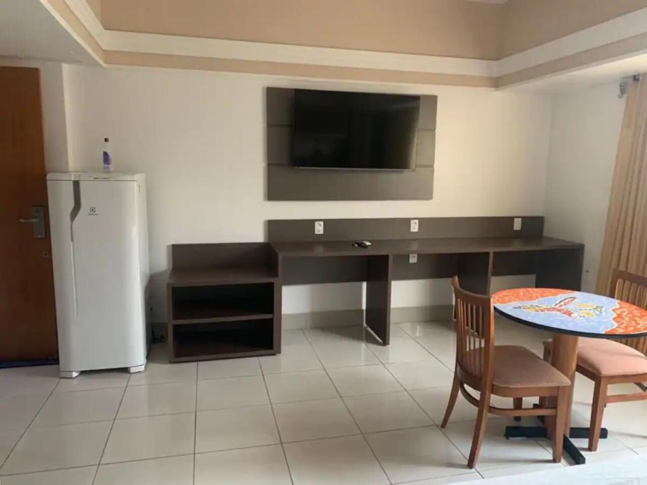 A compact living space with a kitchenette, a flat-screen TV mounted on the wall, a small dining table with chairs, and a tiled floor.