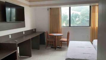 A hotel room with a bed, a desk, a chair, a TV mounted on the wall, and a window with curtains.