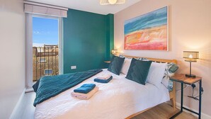 Ein Schlafzimmer mit Bett, Kissen, einem Nachttisch mit Lampe, einem Bild an der Wand und einem Fenster mit Aussicht.