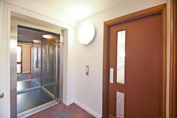 A hallway with a wooden door, a glass elevator door, and a light fixture on the wall.