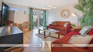 Ein modernes Wohnzimmer mit einer Couch, Couchtisch und Fernseher. Ein großes Fenster mit Vorhängen bietet Blick auf umliegende Gebäude.