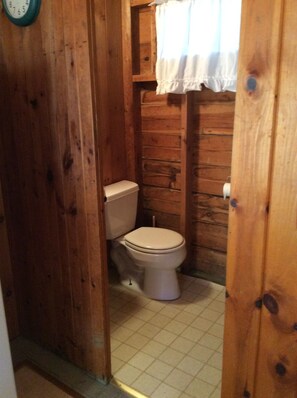 Un bany de fusta amb vàter, una paret de fusta i una finestra amb cortines blanques.