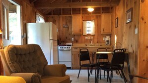 A cozy cabin interior with a dining area, a kitchen with wooden walls, a white refrigerator, and a large cushioned chair. - Brookside Cottage-Private Beach-Pets Welcome - Round Table Lodge, Sebago, Maine (Sebago)