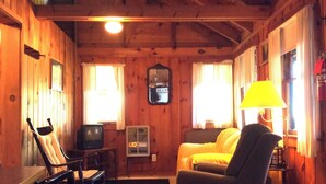 A cozy cabin interior with wooden walls, a bed, a chair, a table, a television, and a lamp. - Brookside Cottage-Private Beach-Pets Welcome - Round Table Lodge, Sebago, Maine (Sebago)