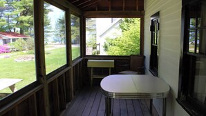 Un porche cubierto con mesa y sillas, ventanas amplias y vista a un jardín y casas vecinas.