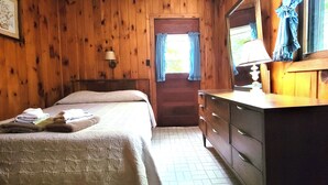 Quarto de madeira com uma cama, cômoda e porta que dá para o exterior.