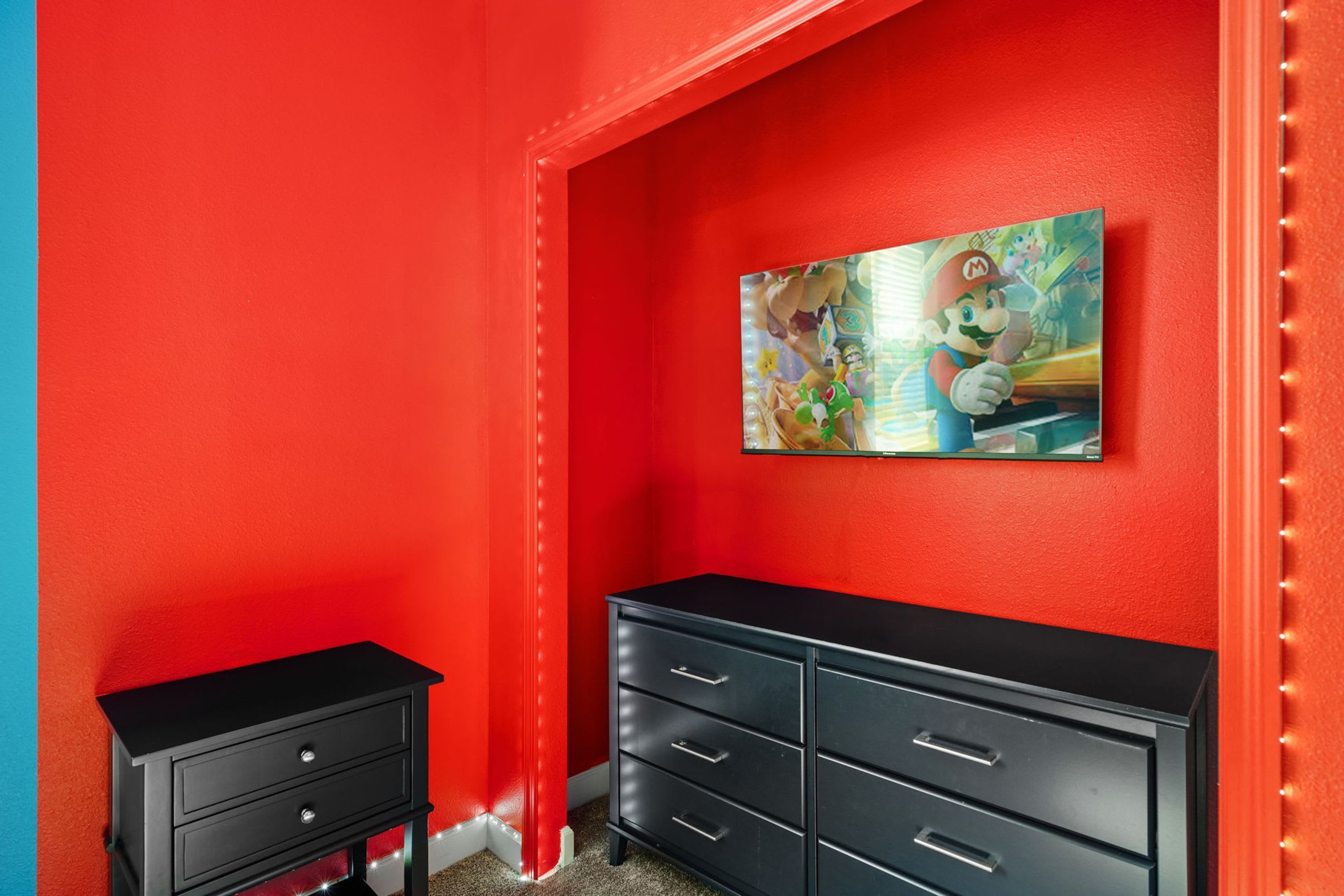 Quarto com paredes vermelhas, um aparador preto e uma pintura com tema do Mario na parede.