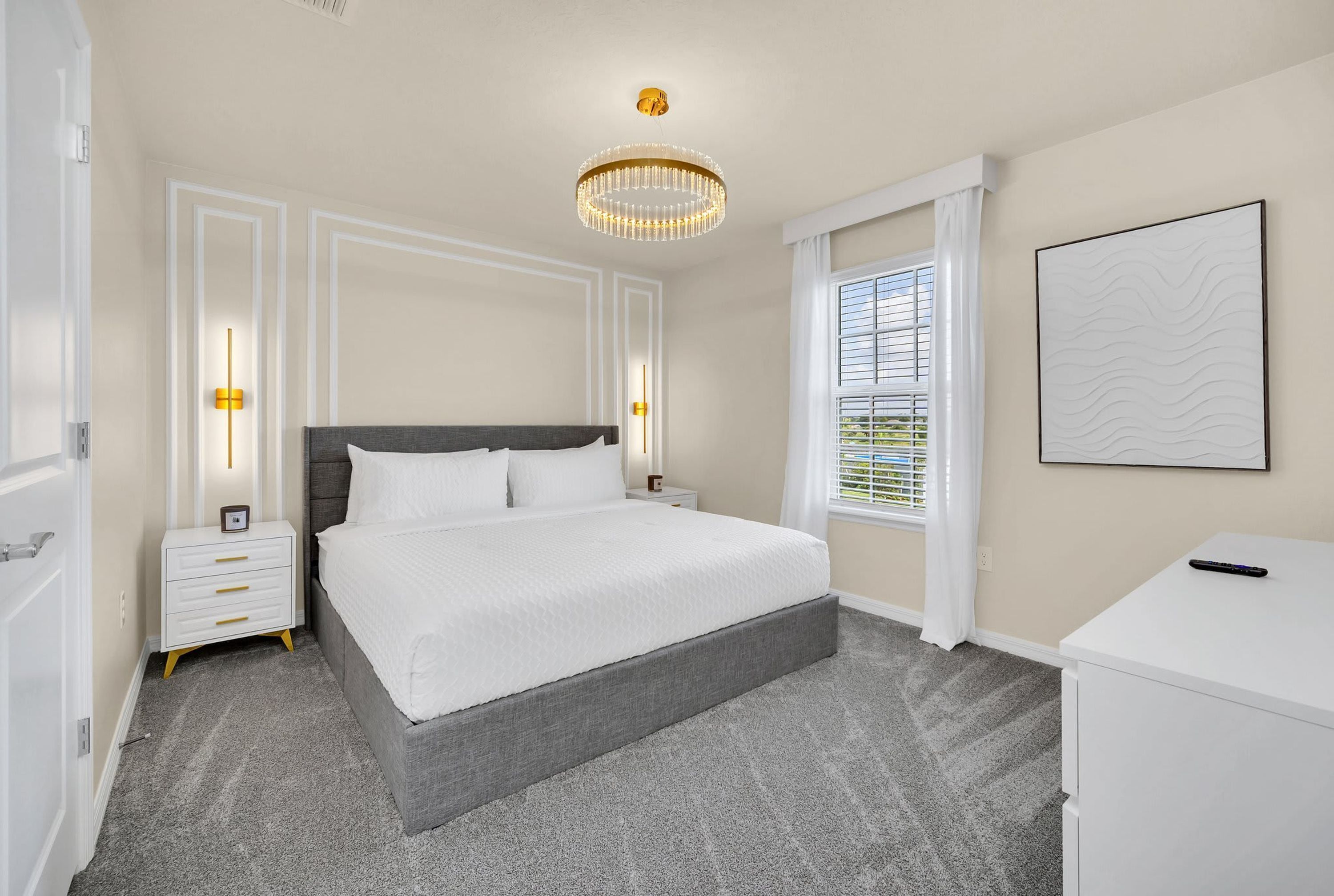 A bedroom with a large bed, bedside tables, a desk, a chandelier, and a window with blinds.