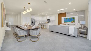 A modern kitchen with white cabinets, a dining area with a wooden table and chairs, and a living area with a sofa and TV.