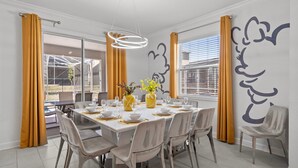 Comedor con una mesa grande dispuesta para seis personas, rodeada de sillas. Hay dos ventanas amplias con cortinas, y una pared con un mural decorativo.
