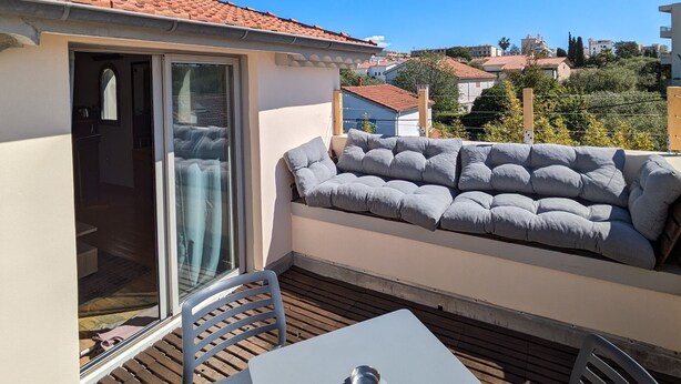 Ein Balkon mit Tisch und Stühlen, eine Couch mit Kissen und eine Glastür, die in einen Innenraum führt.