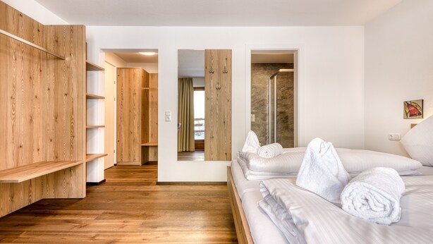 Una habitación de hotel moderna con suelo de madera, una cama grande con ropa blanca, un armario de madera y una zona de ducha.