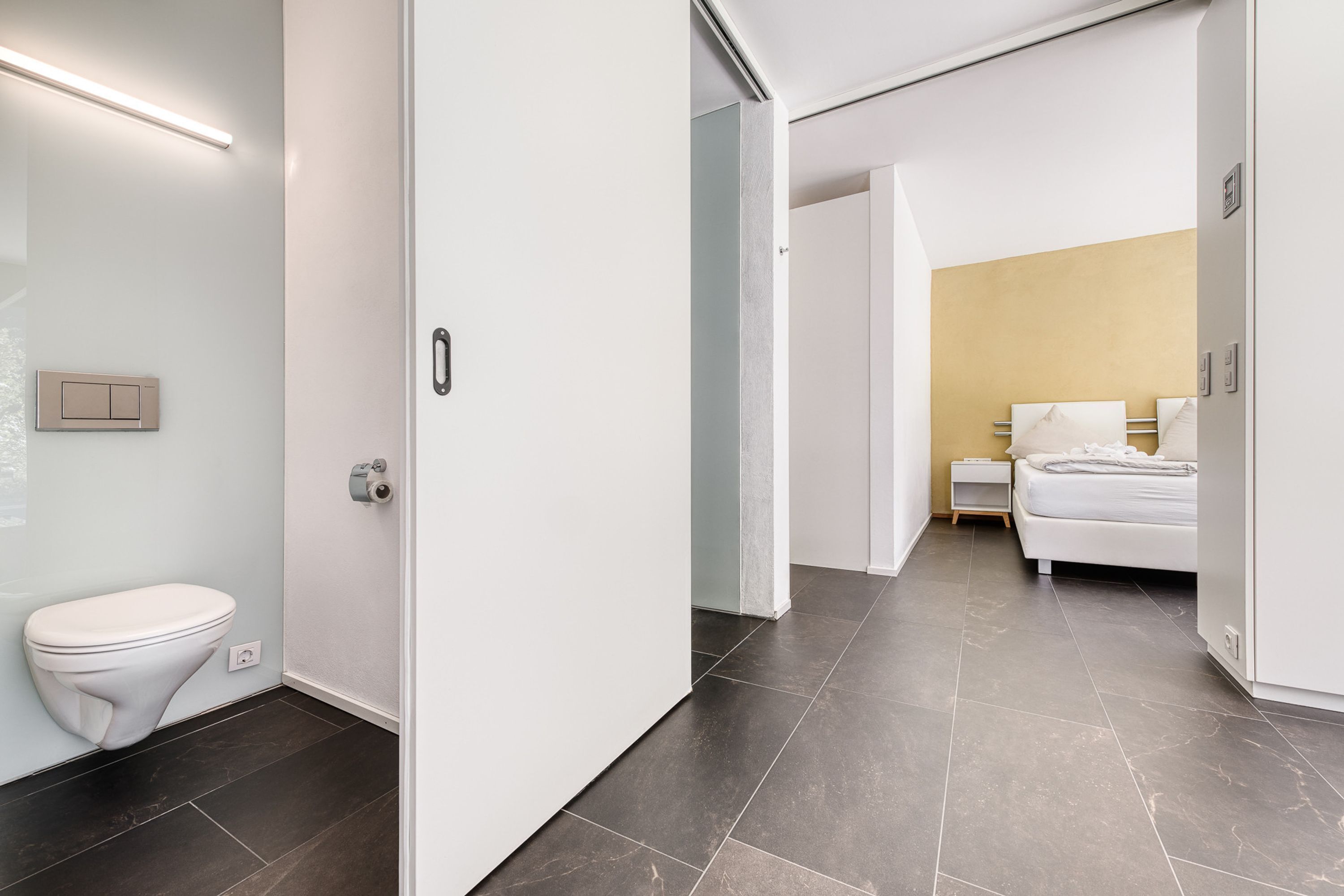 Une salle de bain moderne avec des toilettes blanches, un luminaire fixé au mur et un sol carrelé foncé.