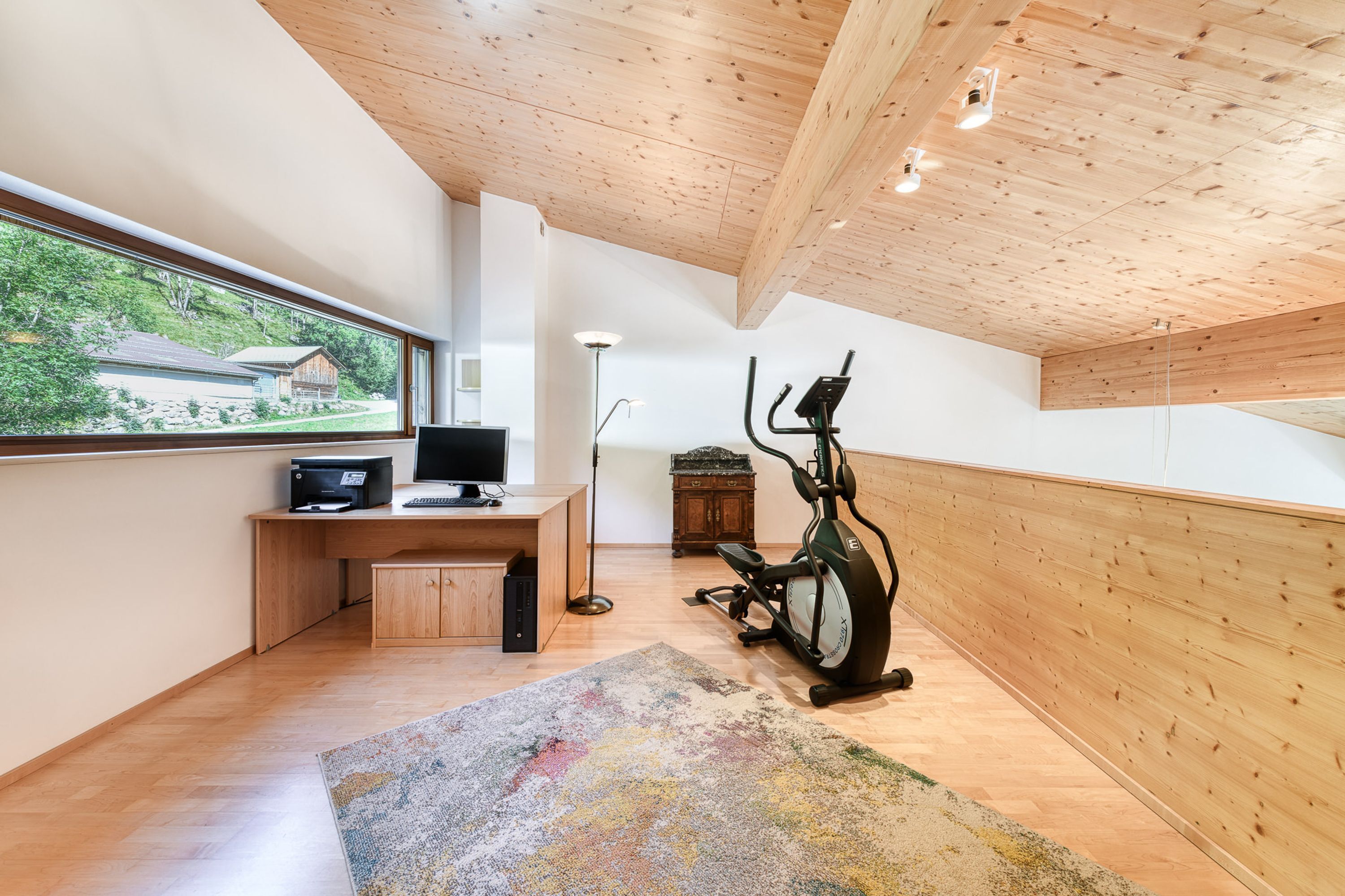 A modern room with a wooden ceiling, a desk with a computer, an exercise bike, and a large window offering a view of a house and greenery.
