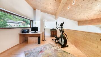 A modern room with a wooden ceiling, a desk with a computer, an exercise bike, and a large window offering a view of a house and greenery.