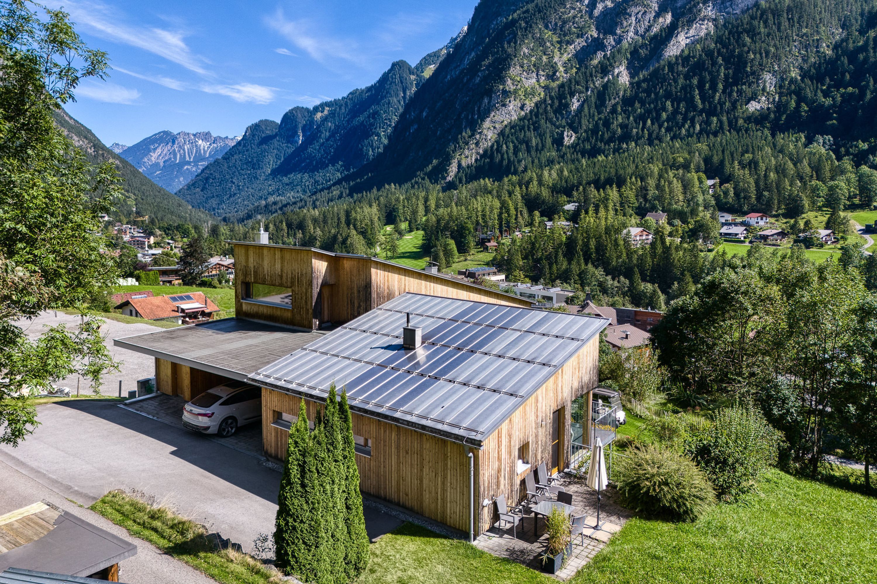 Een modern houten gebouw met een plat dak, een overdekte parkeerplaats en uitzicht op een bergachtig landschap.