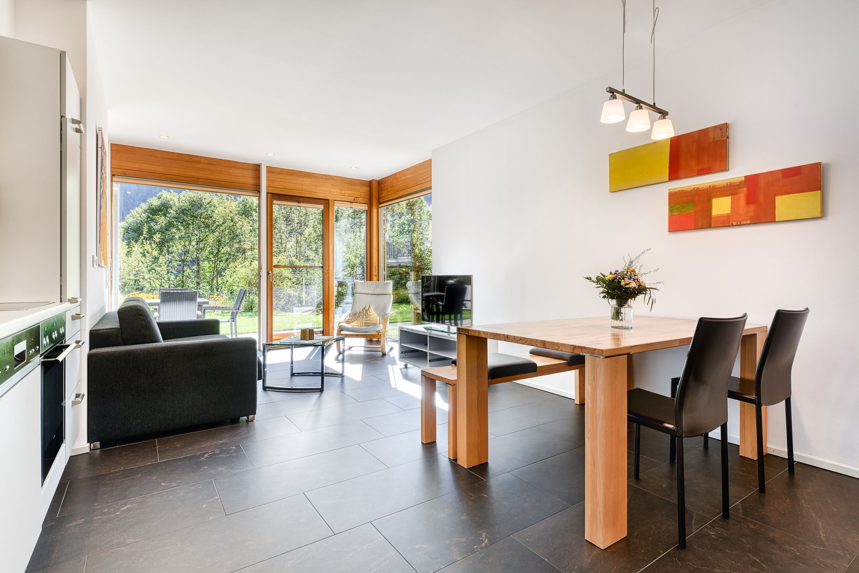 Une cuisine moderne avec des carreaux de couleur sombre, une table à manger en bois et des chaises. On aperçoit un canapé et un téléviseur au fond.