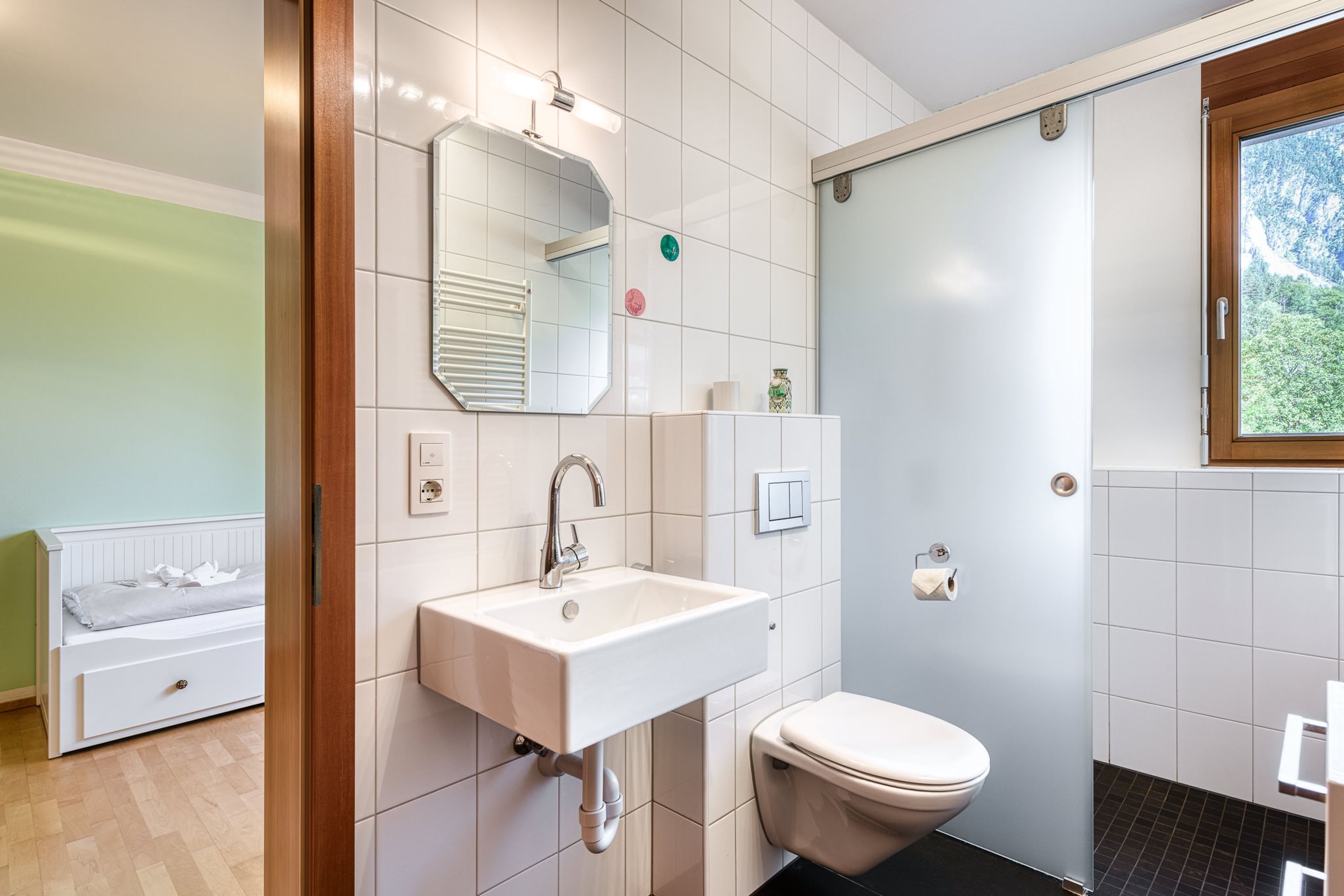 A bathroom with a white sink, a toilet, a mirror, and a window with a view of greenery.