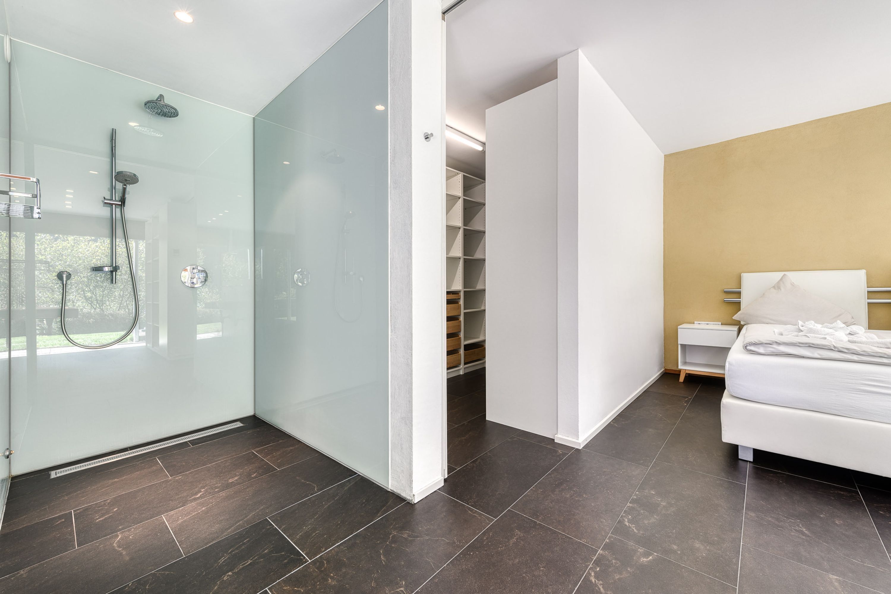 A modern bathroom with a glass-enclosed shower, a white bedside table, and a neatly made bed.
