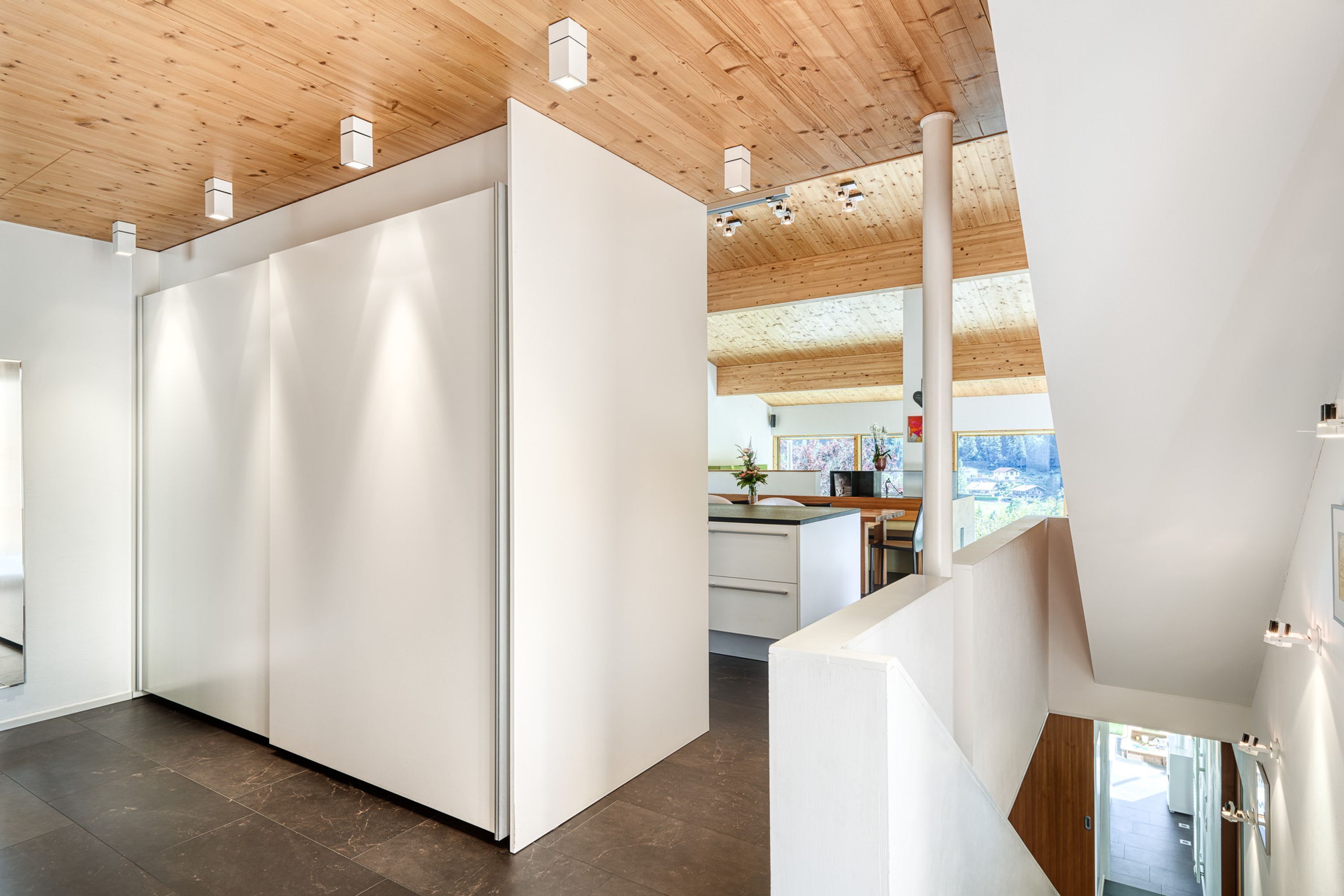Un interior moderno con techo de madera, paredes blancas y una zona de cocina visible a través de una puerta abierta.
