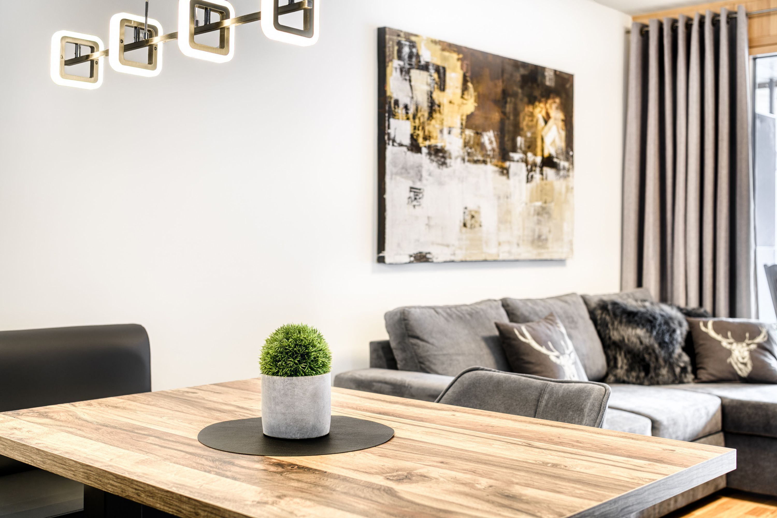 Ein modernes Wohnzimmer mit einem Holzespeisetisch, einem grauen Sofa mit Zierkissen, einem an der Wand hängenden abstrakten Gemälde und einer kleinen Topfpflanze.