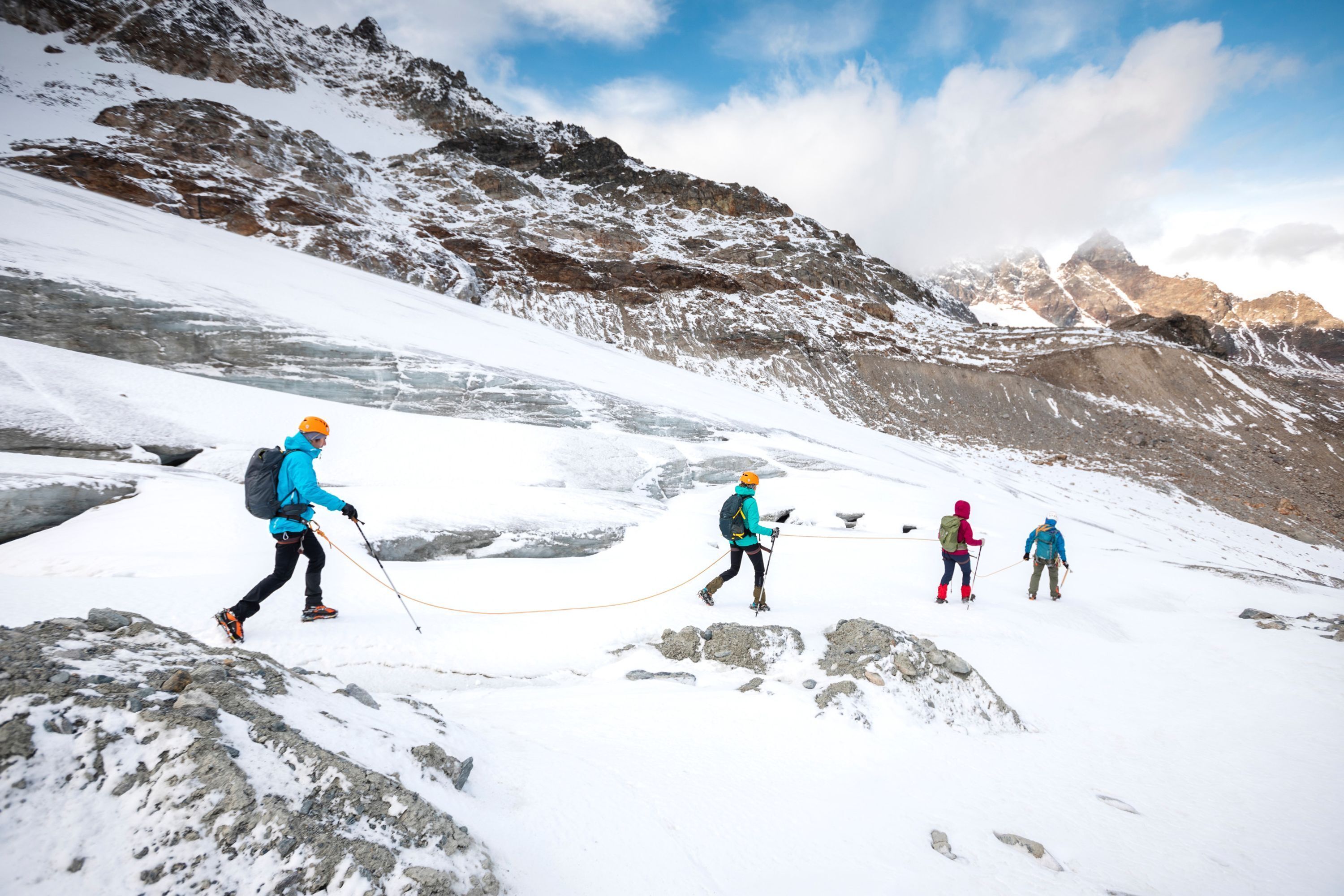 A group of hikers on a snowy mountain trail with rocky terrain and distant peaks.