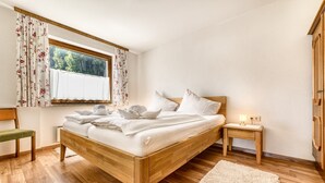 A bedroom with a wooden bed, a nightstand, a bench, a window with curtains, and a view of trees. - Appartement Schruns - M Ser-apartments (Tschagguns)