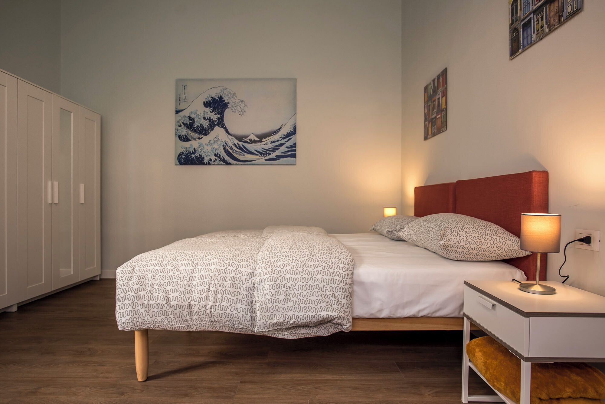 Une chambre à coucher avec un lit, une table de chevet, une lampe et une œuvre d’art accrochée au mur.