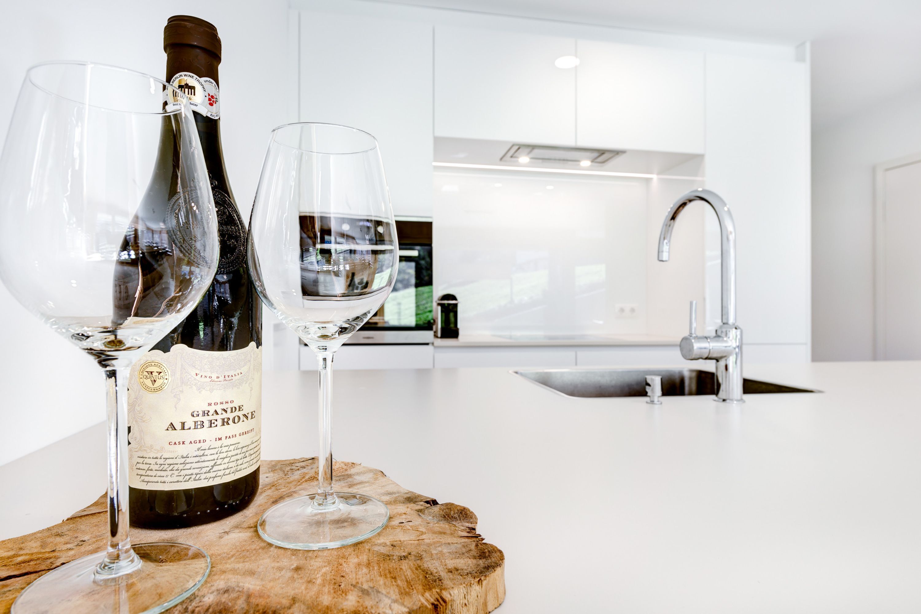 A bottle of wine and two glasses on a wooden board in a modern kitchen with white cabinets and a stainless steel sink.