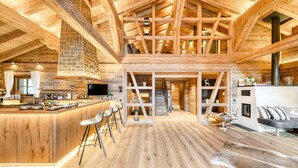 A spacious, high-ceilinged wooden interior with a central bar area, seating, and a staircase leading to an upper level. - Chalet GM - Penthaus (Brand)