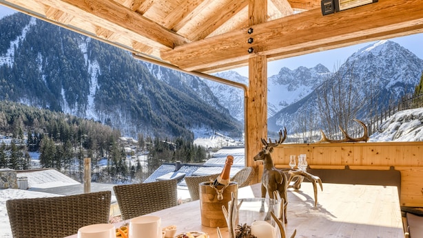 A wooden dining table with a view of snow-covered mountains, a bottle of champagne, and a deer head decoration. - Chalet GM - Penthaus (Brand)