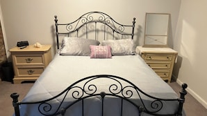 Ein ordentlich bezogenes Bett mit Metallkopfteil, zwei Nachttischen und einem Spiegel an der Wand.