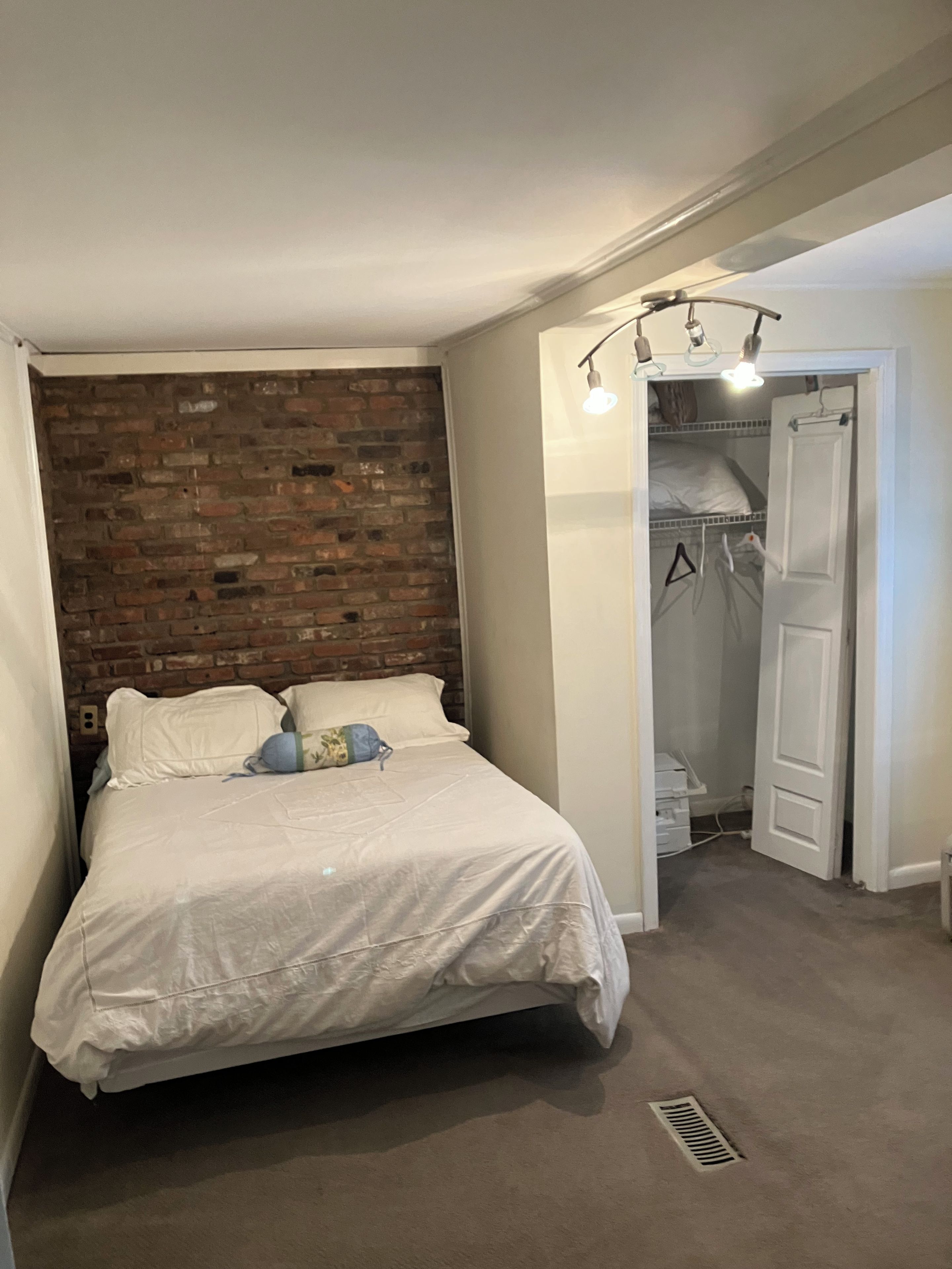 A bedroom with a brick accent wall, a bed with white bedding, a closet with hanging clothes, and a door leading to another room.