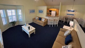 A spacious living area with a high ceiling, a ceiling fan, a kitchen with wooden cabinets, a dining area with bar stools, and a large window with blinds.