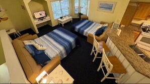 A room with two beds, a desk, a chair, a ceiling fan, a window, and a kitchen area.