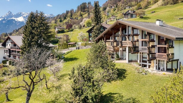 Eine Berghütte mit mehreren Balkonen, umgeben von üppiger Vegetation und einer Kulisse schneebedeckter Berge.