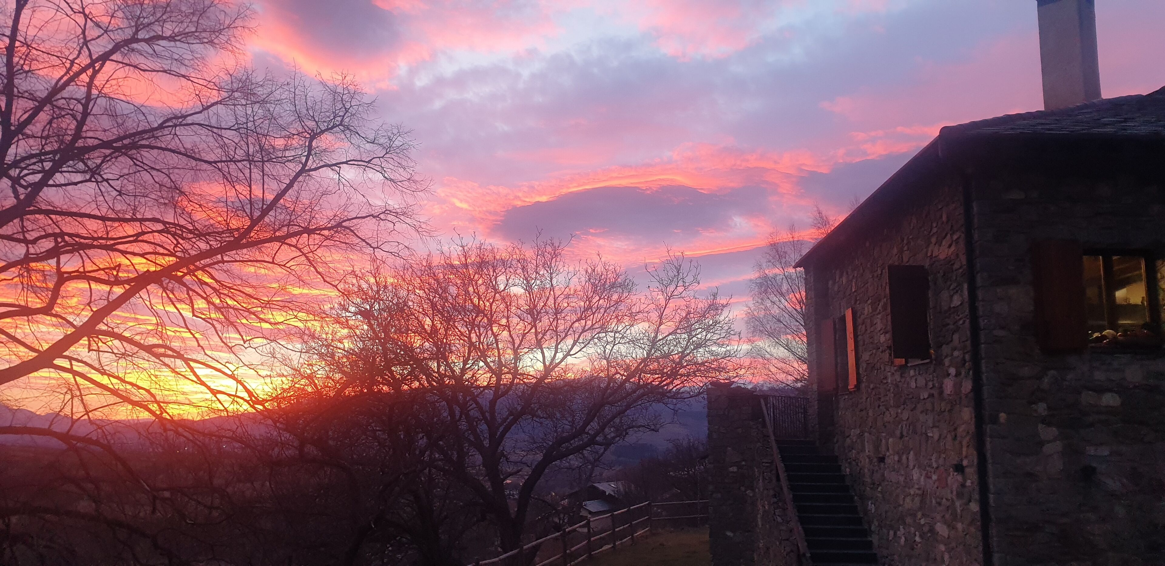 Ein rustikales Steinhaus mit einer Treppe, die zu einem Fenster hinaufführt, vor einer Kulisse aus Sonnenuntergang mit rosa und orangefarbenen Tönen.