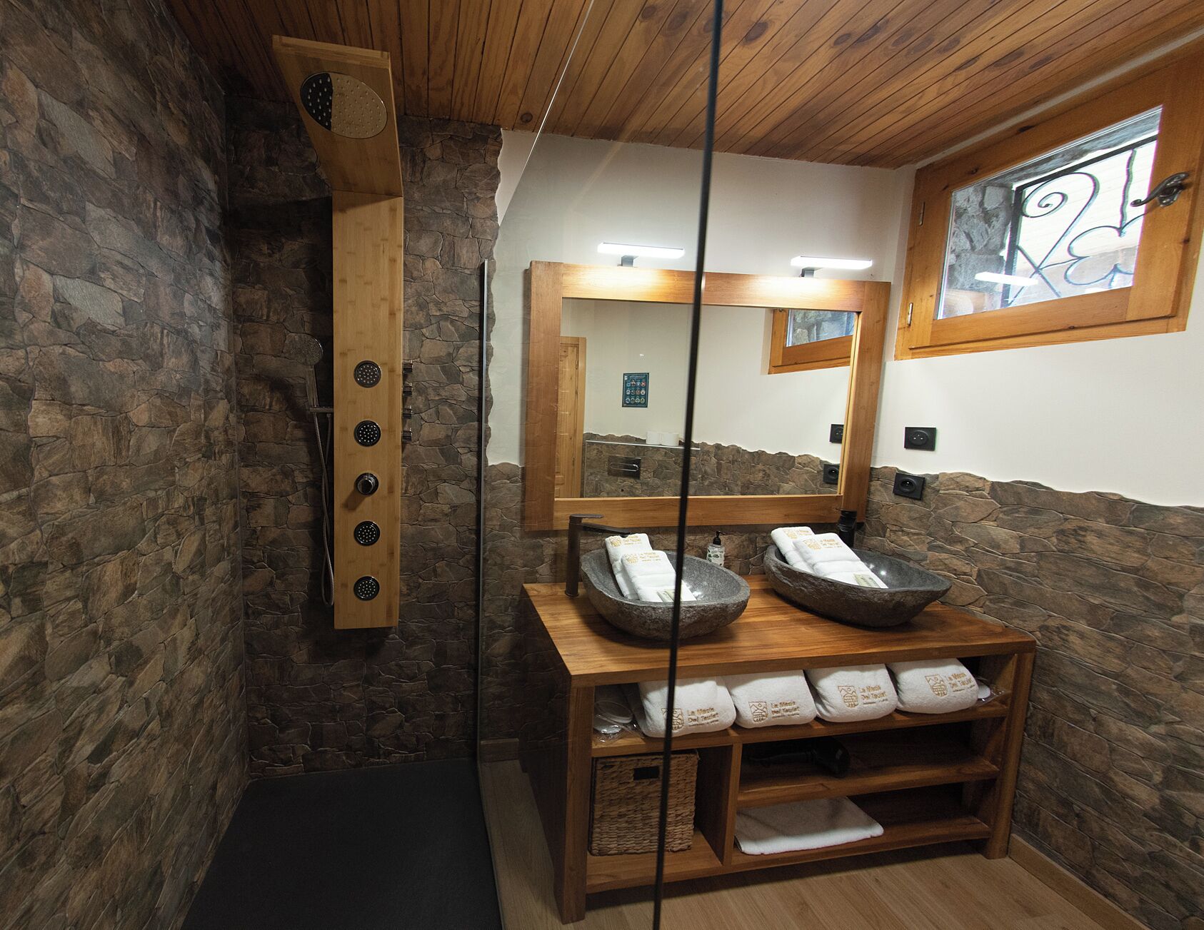 A stone-walled bathroom with a wooden shower enclosure, a large mirror, and a wooden vanity with towels.
