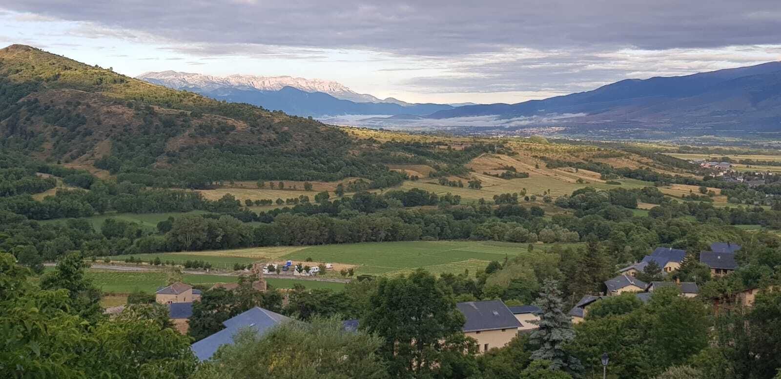 Un paysage rural caractérisé par des collines douces, des maisons éparpillées et une chaîne de montagnes qui se dessine à l’horizon.
